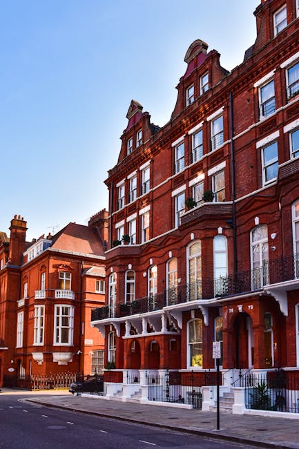 Photograph of a multi-storey red brick residential building on a city street, featuring large sash windows and decorative iron balcony railings on the lower floors. The building's ground floor has an 