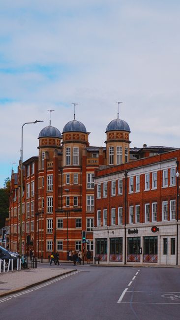 Photograph of a multi-storey red brick residential building on a city street, featuring large sash windows and decorative iron balcony railings on the lower floors. The building's ground floor has an 