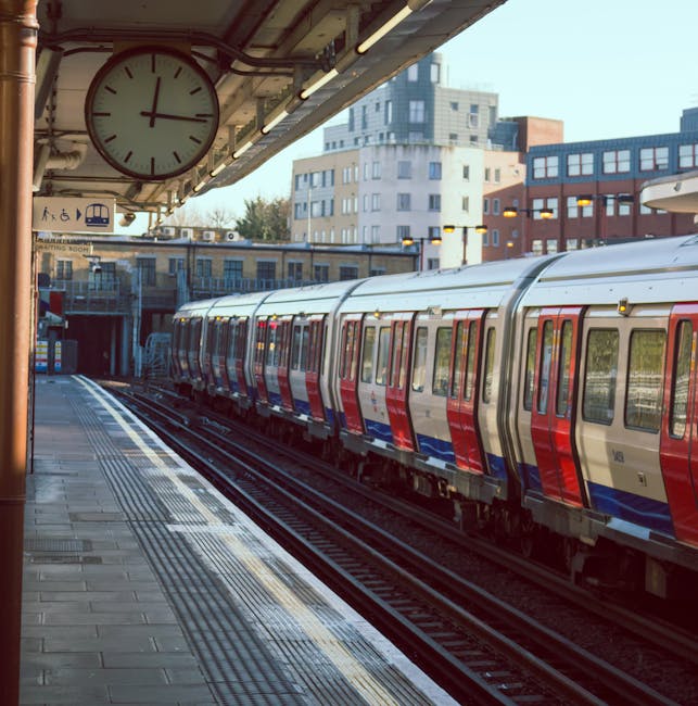 A modern London Underground train with a silver body and red and blue accents parked at an outdoor station platform. The platform is paved with grey tiles and has tactile paving strips along the edge for safety. Overhead, a large analog clock with a white face, black minute and hour hands, and a red second hand is mounted under the station's concrete canopy. The canopy is equipped with fluorescent lighting and visible piping. In the background, contemporary residential and commercial buildings with varied window styles and brick or concrete facades can be seen, indicating an urban environment near Clapham Junction station. The scene captures the typical setting for a home relocation or moving process, emphasizing the importance of efficient transport logistics and the availability of nearby train services, as managed by Removal Companies Wandsworth.