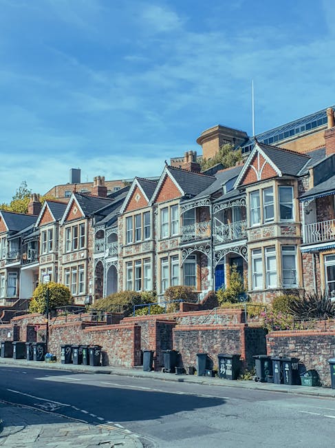 A row of Victorian-style terraced houses in Wandsworth with brick facades, decorative white balcony railings, and bay windows facing the street. In the foreground, a pavement lined with black wheelie bins and recycling containers is visible. Outside one of the houses, a moveable dolly with a large, partially wrapped cardboard box and a plastic cover for furniture is positioned near the doorway, indicating a home relocation process. Bright daylight illuminates the scene, with a partly cloudy blue sky overhead. The environment is quiet, and the image captures a typical residential street preparing for or in the process of house removals, with no moving personnel visible but equipment suggesting ongoing packing or furniture transport managed by local removals service providers, such as Removal Companies Wandsworth.